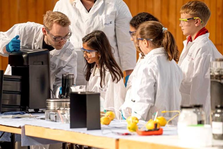 Eine Gruppe von Kindern und ein Erwachsener in Laborkitteln und Schutzbrillen an einem Experimentiertisch