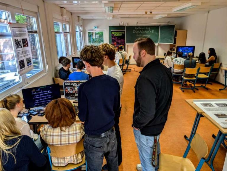 Menschen in einem Klassenraum betrachten mehrere Computerbildschirme und Ausstellungstafeln zu Retrospielkonsolen.