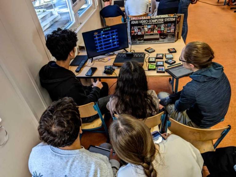 Mehrere Personen sitzen um einen Tisch und sehen sich eine Atari-Spielkonsole mit Bildschirm an.