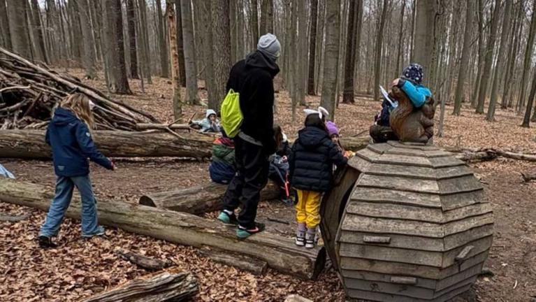 Kinder und eine erwachsene Person im Wald neben einer großen Holzskulptur und umgestürzten Baumstämmen