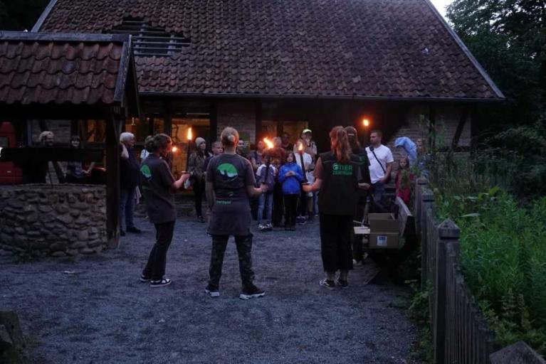 Eine Gruppe von Menschen steht bei Dämmerung vor einem Haus und hält brennende Fackeln.