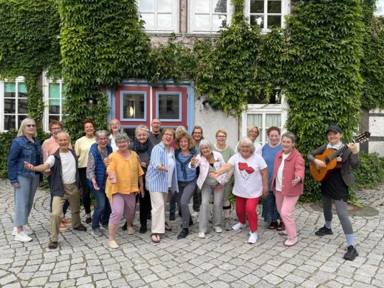 Eine Gruppe von Frauen und einem Mann steht lachend vor einem efeubewachsenen Gebäude, rechts spielt eine Frau Gitarre.