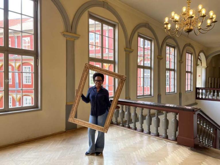 Eine Person hält einen großen leeren Bilderrahmen in einem hellen Treppenhaus mit hohen Fenstern und Kronleuchter.