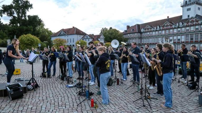 Eine Blaskapelle spielt auf einem gepflasterten Platz im Freien vor historischen Gebäuden, dirigiert von einer Frau.