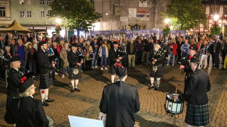 Eine Dudelsackgruppe in schottischen Uniformen spielt auf einem Platz vor einer Menschenmenge bei Nacht.