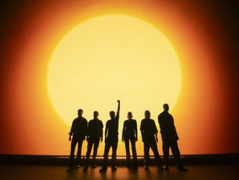 Sechs Menschen stehen als Silhouetten vor einer riesigen leuchtenden Sonne.