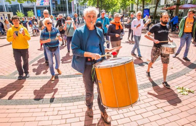 Mehrere Menschen spielen und laufen in einer sonnigen Fußgängerzone bei einer Straßenmusikaktion, im Vordergrund ein Mann mit großer Trommel.