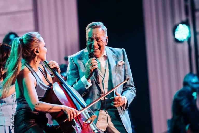 Ein älterer Mann singt mit Mikrofon neben einer Cellistin auf einer Bühne.