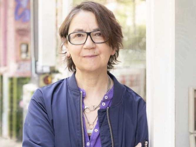 Porträt einer Frau mit Brille und dunkler Jacke vor unscharfem Hintergrund