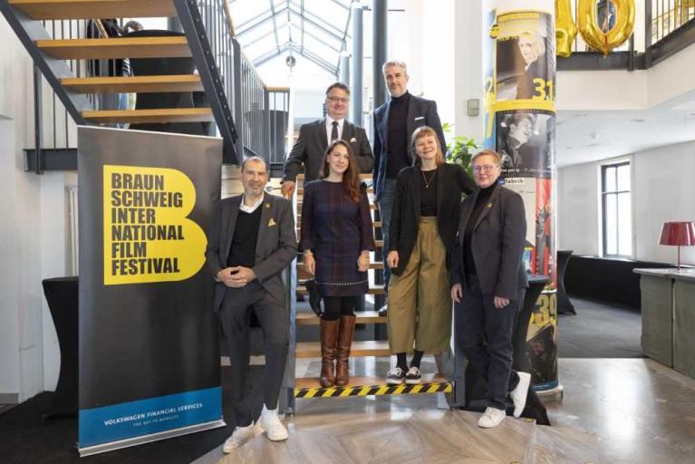 Sechs Personen posieren auf einer Treppe neben einem Banner des Braunschweig International Film Festival.