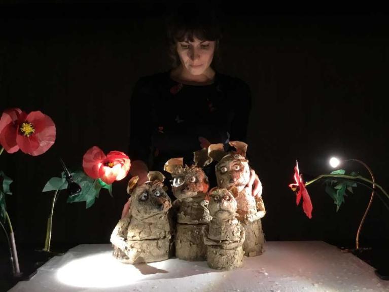 Eine Frau steht hinter vier handgefertigten Tierfiguren auf einem Tisch, flankiert von leuchtenden Papierblumen.
