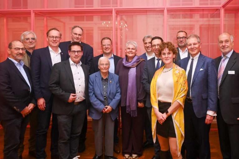 Gruppe von zwölf formal gekleideten Personen posiert vor einer roten Glasfassade