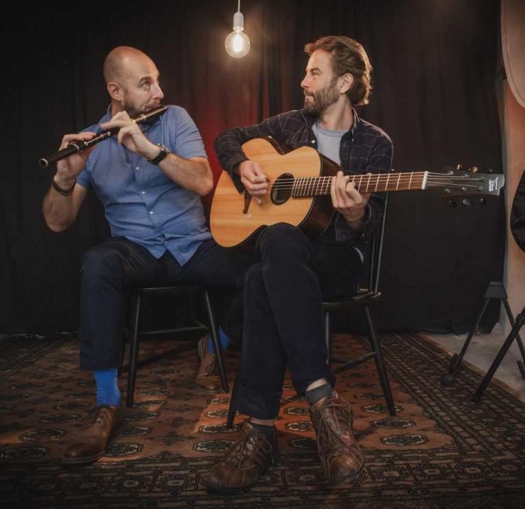 Zwei Männer sitzen auf Stühlen und spielen Flöte und Gitarre vor dunklem Hintergrund.