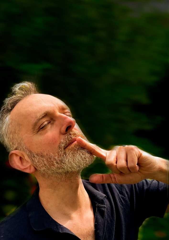 Ein bärtiger Mann mit geschlossen Augen berührt mit dem Finger sein Kinn vor grünem Hintergrund.