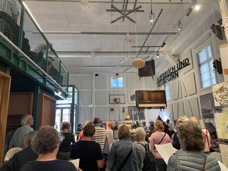Eine Gruppe von Besuchern steht in einem hohen Ausstellungsraum und blickt auf hängende Objekte und Wandtexte.