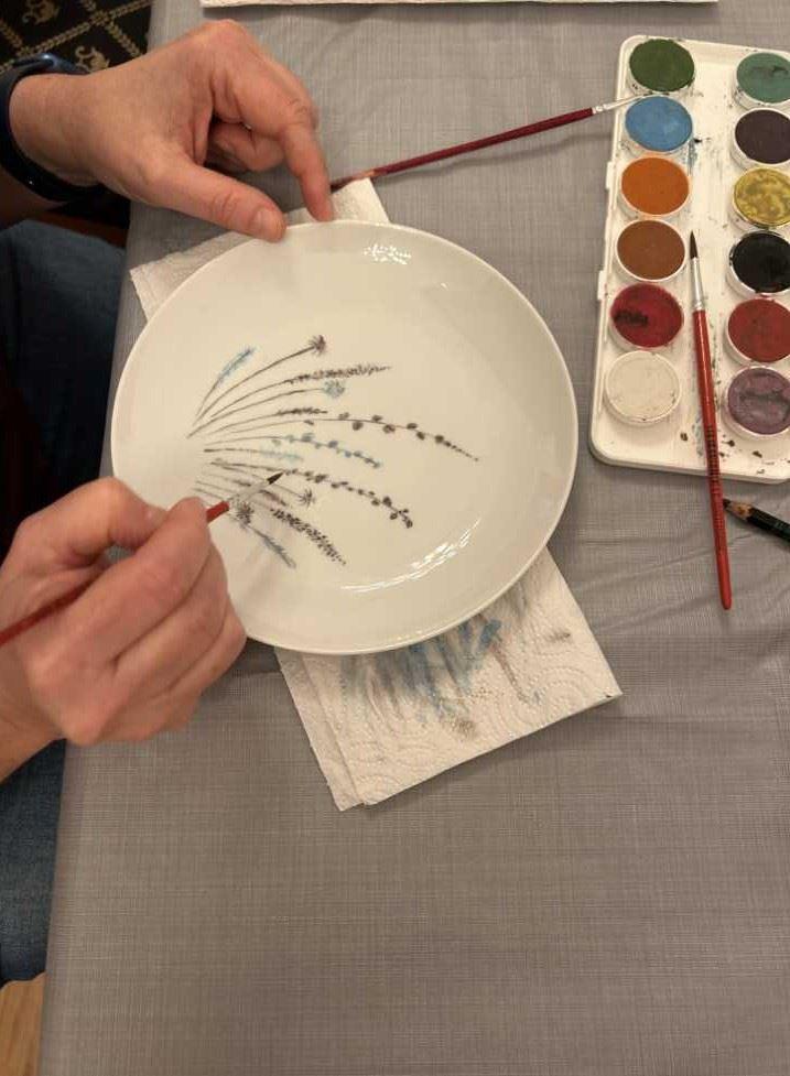 Hände bemalen einen weißen Teller mit feinen Pflanzenmotiven neben einer Farbpalette