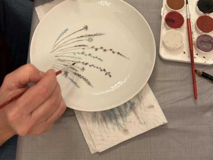 Hände bemalen einen weißen Teller mit feinen Pflanzenmotiven neben einer Farbpalette