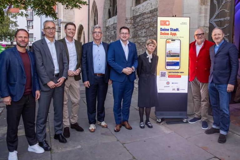 Mehrere Personen stehen vor einem historischen Gebäude neben einem digitalen Werbedisplay für eine App.