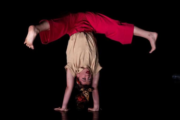 Eine Person hängt kopfüber in einem Handstand mit gespreizten Beinen vor schwarzem Hintergrund.