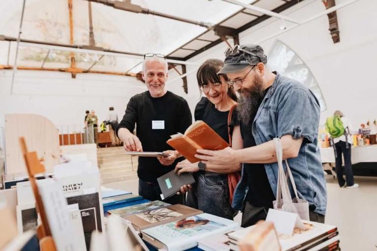 Drei Personen betrachten Bücher an einem Büchertisch in einer hellen Halle