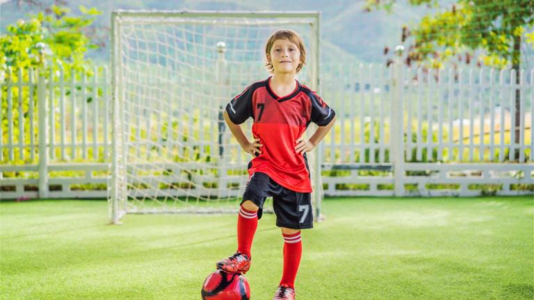 Junge im roten Fußballtrikot steht mit einem Fuß auf einem Fußball vor einem Tor