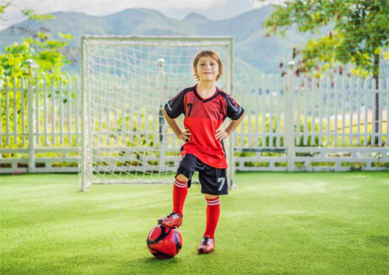 Junge im roten Fußballtrikot steht mit einem Fuß auf einem Fußball vor einem Tor
