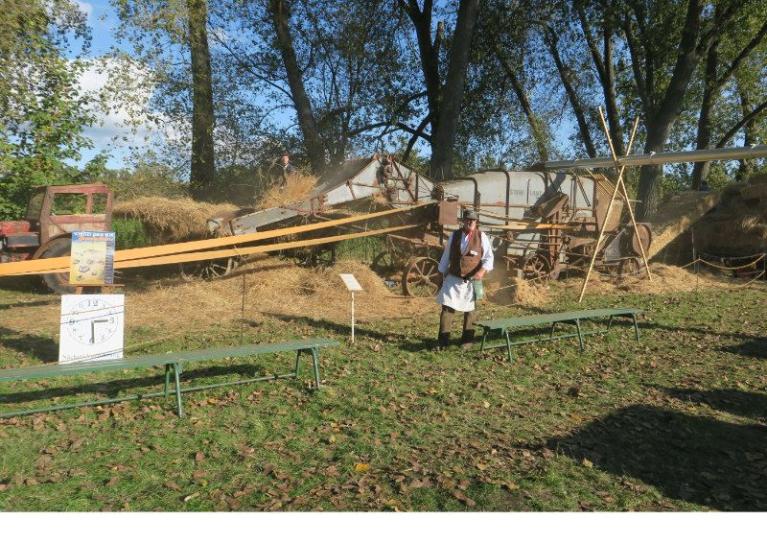 Eine historische landwirtschaftliche Maschine im Freien, davor steht eine Person in traditioneller Kleidung.