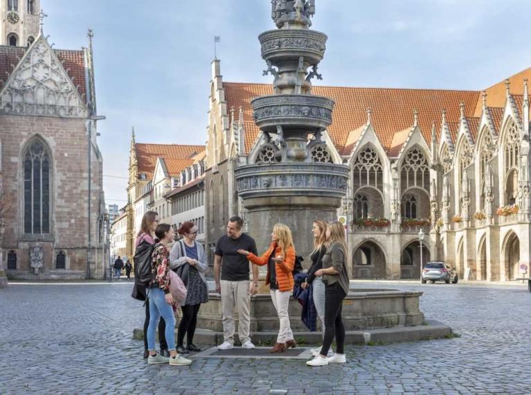 Gruppe von Menschen steht auf einem historischen Platz vor einem steinernen Brunnen und gotischen Gebäuden