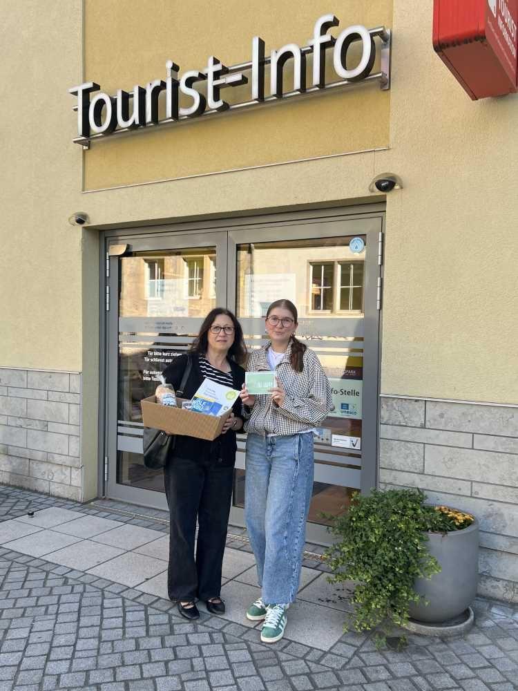 Zwei Frauen stehen vor einer Tourist-Info und halten Werbematerial und eine Box mit Infomaterial