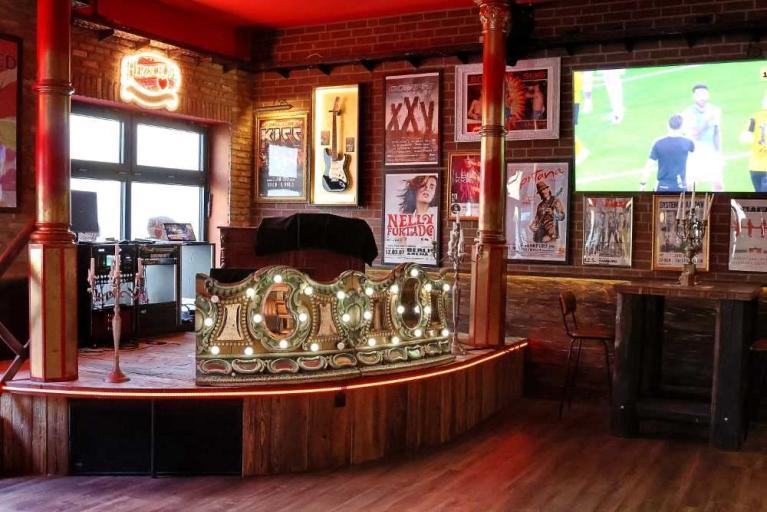 Innenraum einer Bar mit Bühne, Neonlicht, Wandpostern und Fernseher mit Sportübertragung