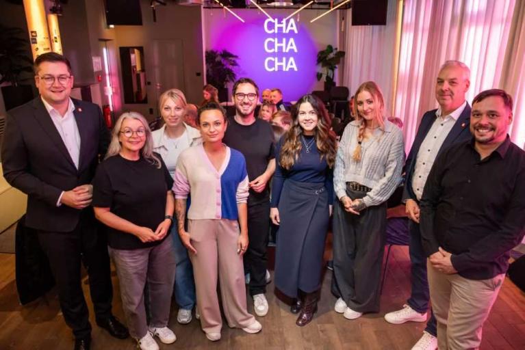 Gruppe von neun Personen steht bei einer Veranstaltung in einem beleuchteten Raum vor einer Bühne mit dem Schriftzug CHA CHA CHA.