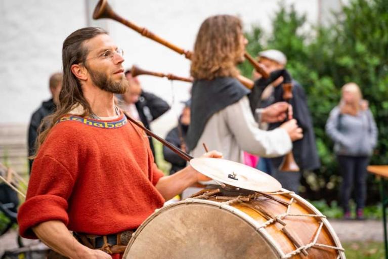 Ein Mann in rotem Pullover spielt ein großes Trommelinstrument, im Hintergrund stehen Personen mit Blasinstrumenten.
