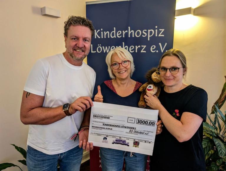 Drei Personen halten einen Spendenscheck vor einem Banner des Kinderhospiz Löwenherz e.V.