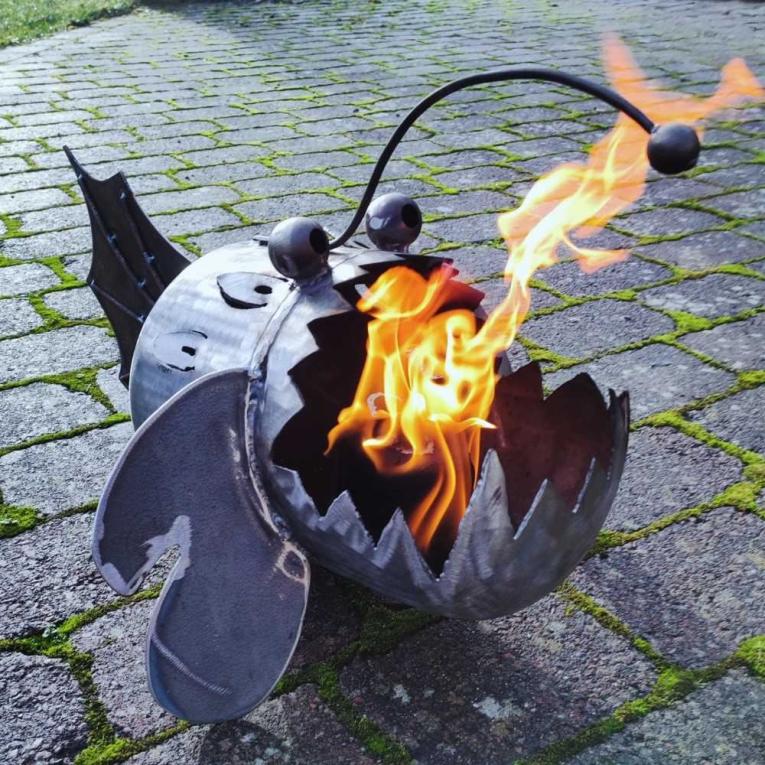 Ein metallisches Feuerobjekt in Form eines Fisches brennt auf gepflastertem Boden.