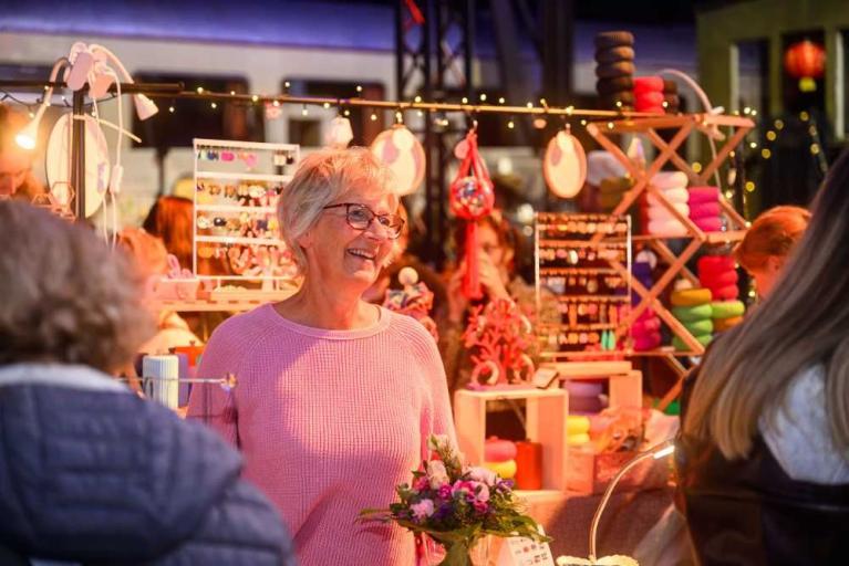 Lächelnde ältere Frau in pinkem Pullover an einem Marktstand mit buntem Schmuck und Accessoires