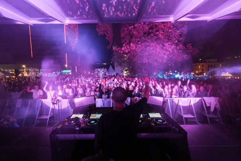 DJ von hinten vor einer großen Menschenmenge bei einem nächtlichen Konzert mit violetter Beleuchtung