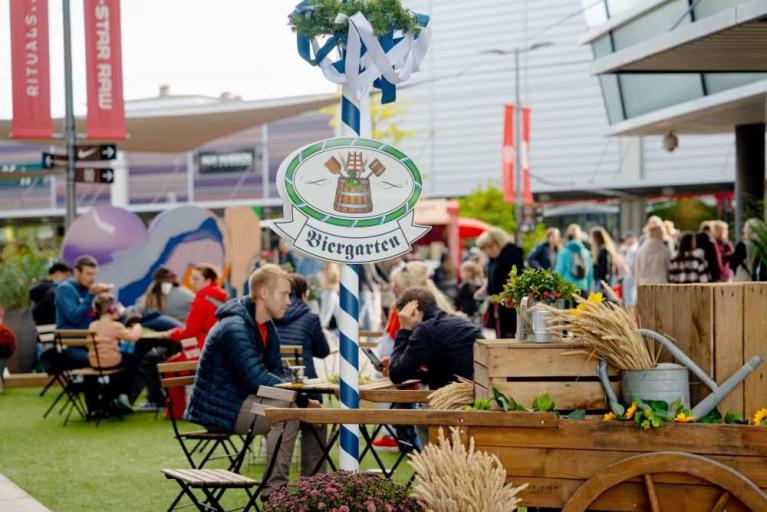 Biergarten mit Sitzplätzen im Freien und dekoriertem Schild in einer belebten Umgebung