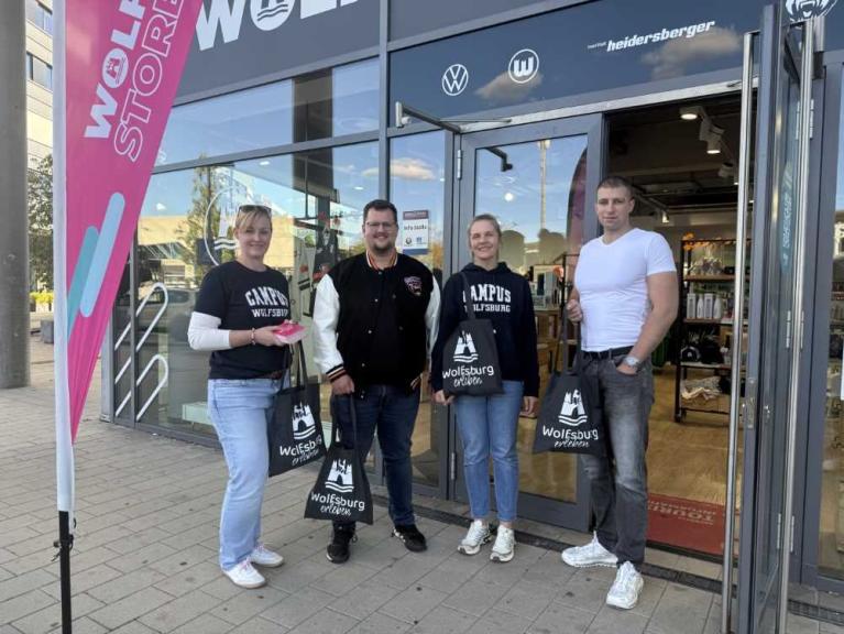 Vier Personen stehen vor einem Laden mit Wolfsburg- und VW-Logo und halten Einkaufstaschen