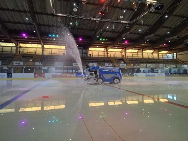 Eine blaue Reinigungsmaschine sprüht Wasser auf eine Eisfläche in einer Eishalle