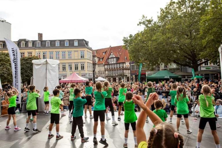 Kinder in grünen Shirts tanzen auf einer Bühne vor einer Menschenmenge auf einem Stadtplatz