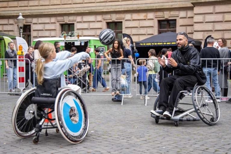 Zwei Personen im Rollstuhl spielen vor einer Menschenmenge auf einem Platz