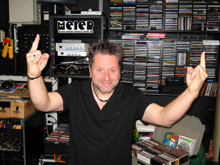 Ein Mann in schwarzem Shirt steht in einem Raum voller CDs und hebt beide Zeigefinger.
