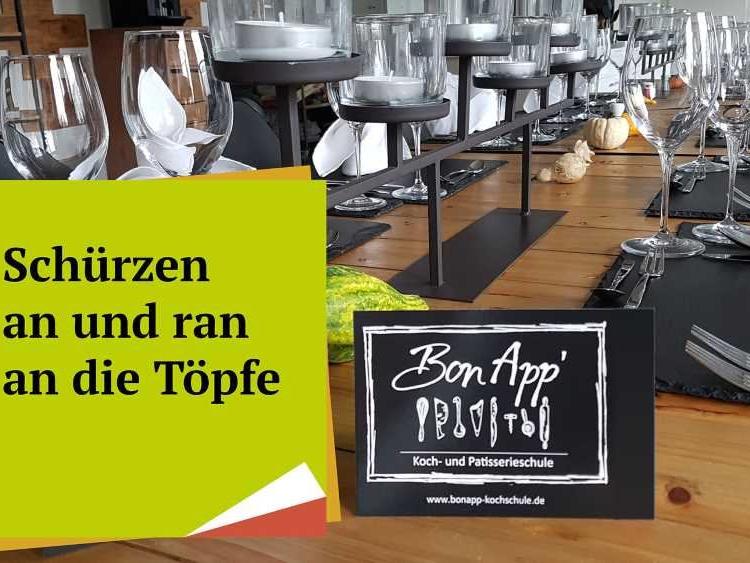 Festlich gedeckter Holztisch mit Gläsern, Besteck und Tischkarten, davor ein Schild mit BonApp und ein grünes Plakat mit Text