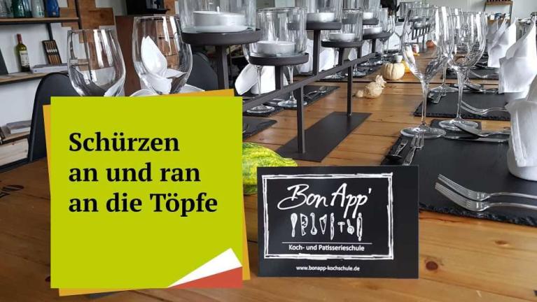 Festlich gedeckter Holztisch mit Gläsern, Besteck und Tischkarten, davor ein Schild mit BonApp und ein grünes Plakat mit Text