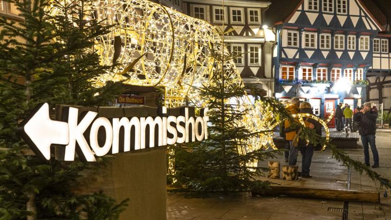 Beleuchtetes Kommissor-Schild auf einem festlich dekorierten Platz mit Weihnachtsbeleuchtung und Fachwerkhäusern