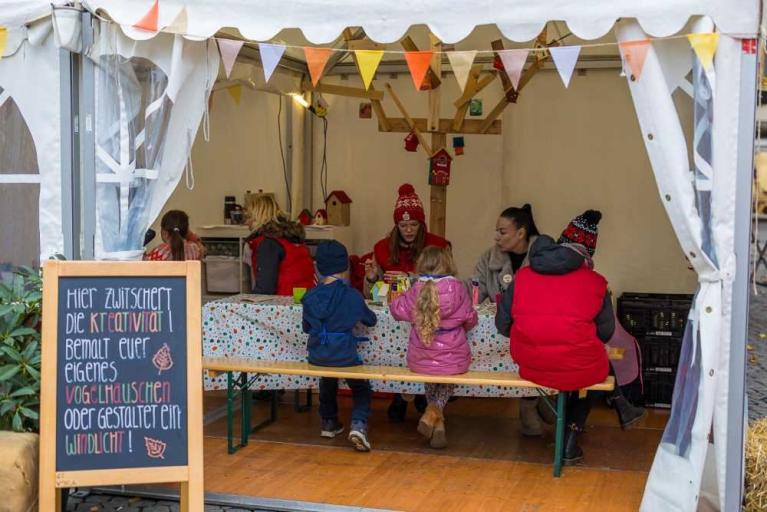 Kinder sitzen an einem Basteltisch in einem Zelt mit einer Kreidetafel im Vordergrund