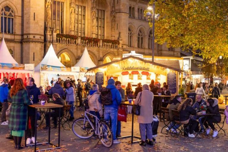 Menschen auf einem beleuchteten Herbstmarkt mit Essensständen vor einem historischen Gebäude