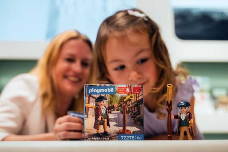 Ein Kind und eine Frau betrachten eine Playmobil-Figur und die Verpackung des Sets Carl Friedrich Gauß.
