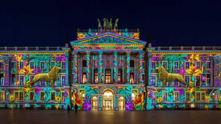 Nachts beleuchtete Fassade eines klassizistischen Gebäudes mit farbenfroher Lichtprojektion und zwei Löwenskulpturen
