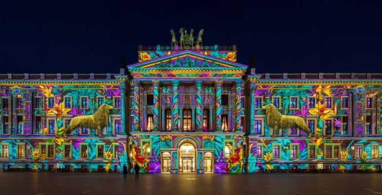 Nachts beleuchtete Fassade eines klassizistischen Gebäudes mit farbenfroher Lichtprojektion und zwei Löwenskulpturen